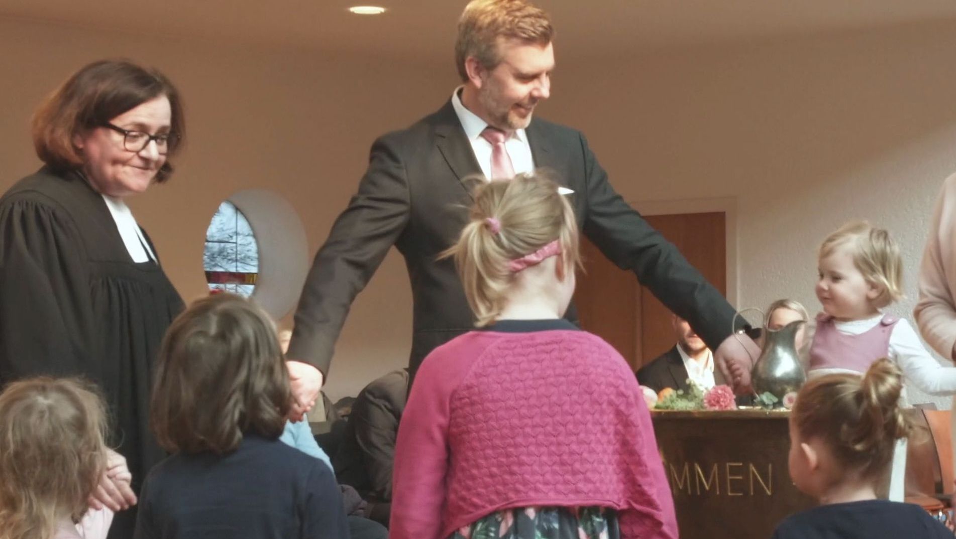 Pfarrerin und Tauffamilie stehen im Kreis um das Taufbecken und fassen sich an der Hand; Foto: Emmaus