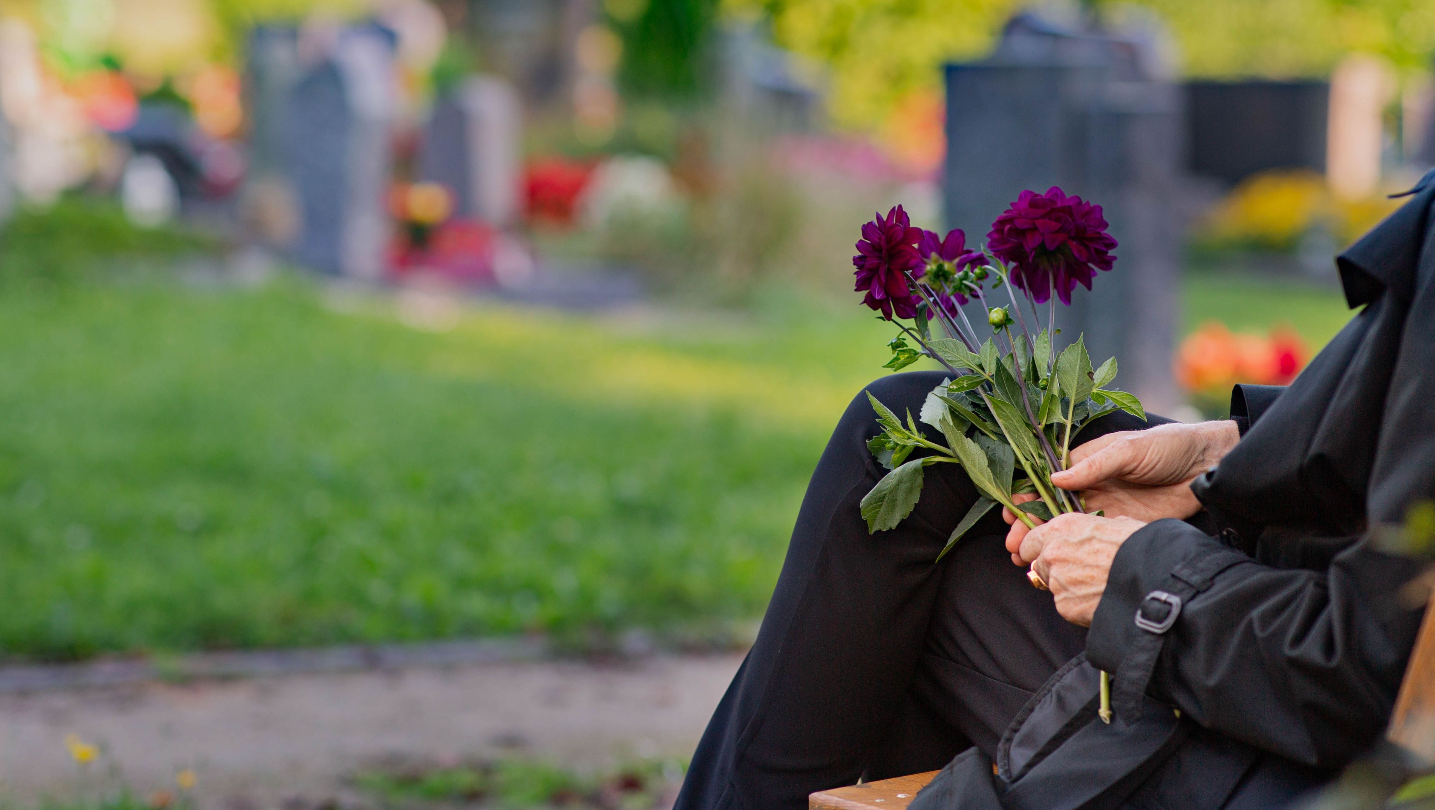Frau sitzt mit Blumen in der Hand auf einer Bank auf dem Friedhof; Foto: Fundus